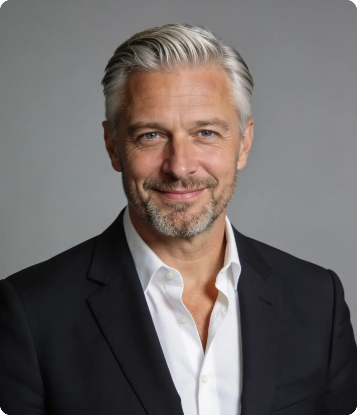 Smiling middle-aged man with gray hair, beard, wearing a black suit and white shirt on a gray background.
