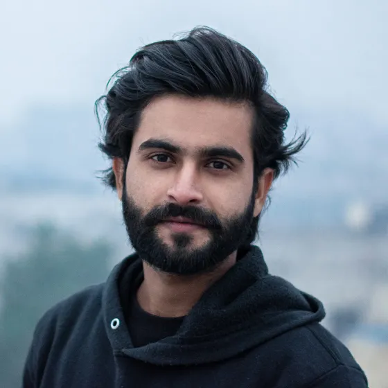 Young man with thick black hair and beard wearing a black hoodie, outdoors with blurred background.