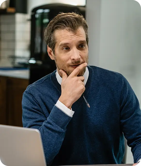 Man in blue sweater sitting at desk, holding pen near chin, looking thoughtfully to the side.