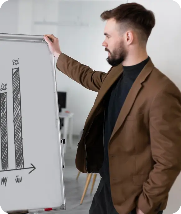 Man in brown jacket pointing at a whiteboard showing a bar graph labeled May and Jun with percentages.