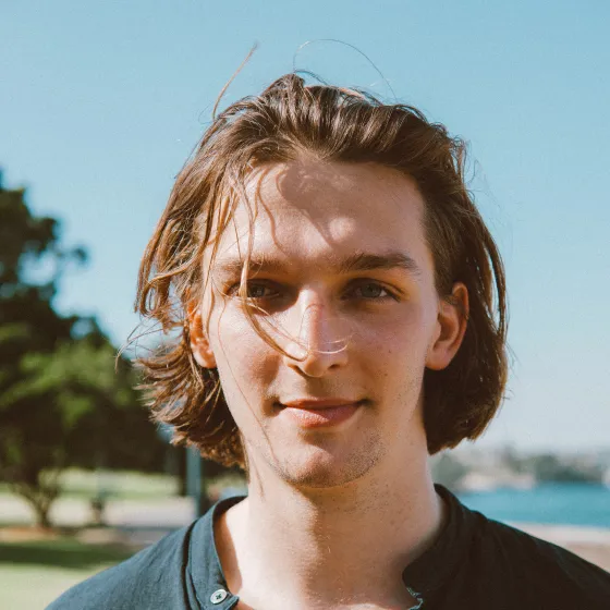 Young man with medium-length hair stands outdoors in sunlight near trees and water.