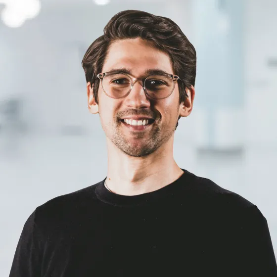 Smiling young man with glasses and wavy brown hair wearing a black shirt in a bright space.